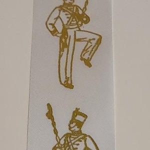 Puede incluir: Cinta blanca con un patrón repetido de ilustraciones doradas de un miembro de una banda de música que sostiene un tambor y un rifle.