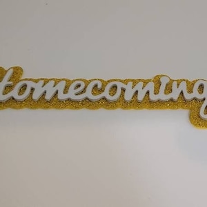 May include: White cursive lettering spelling "Homecoming" on a gold glitter background.