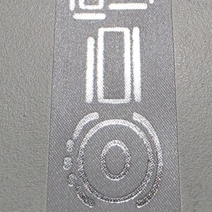 May include: A vertical light gray fabric ribbon featuring the word JUNIOR printed in a metallic silver, stylized font. The letters are arranged in a single column, with each character displaying a distinct, modern, and geometric design.