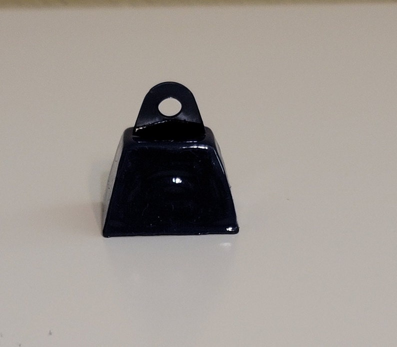 May include: A small, black metal bell with a rounded top and a loop for hanging. The bell has a smooth, shiny finish.