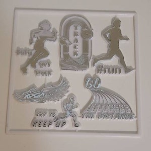 May include: A set of eight silver-colored rubber stamps with track and field themed designs. The stamps feature images of runners, a track, and text such as "Try to keep up", "Go the distance", "Run", and "Get track".