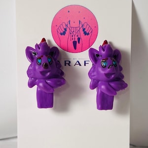 May include: A pair of purple cartoon animal earrings with red accents. The earrings are shaped like a wolf or fox with a fluffy mane and a cute face.