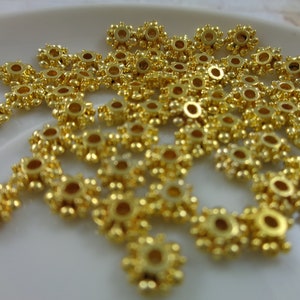 May include: A close-up of a pile of gold-colored metal beads. The beads are round with a small hole in the center and have a decorative design around the edge.