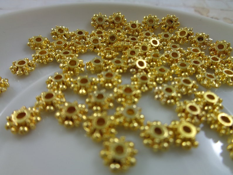 May include: A close-up of a pile of gold-colored metal beads. The beads are round with a small hole in the center and have a decorative design around the edge.