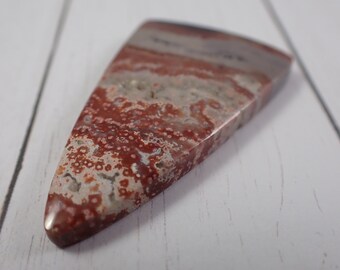 May include: A triangular shaped, red and white agate cabochon with a mottled pattern. The stone has a smooth, polished surface.