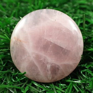 May include: A smooth, round, pink rose quartz crystal with white veins. The crystal is sitting on a bed of green artificial grass.
