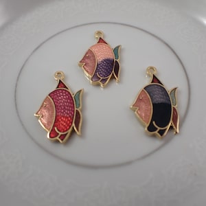 Peut inclure: Trois breloques en forme de poisson colorées avec des bordures dorées. Les poissons ont une variété de couleurs, dont le rose, le rouge, le violet et le noir. Les breloques sont sur une assiette blanche.
