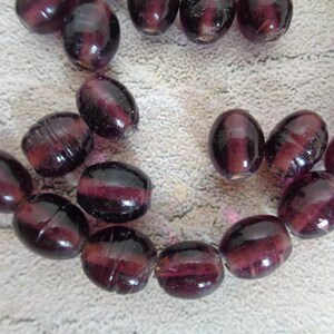 May include: A strand of deep purple glass beads, each bead is oval shaped and has a slight iridescent sheen.