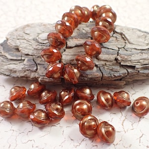 May include: A strand of amber glass beads with a faceted, teardrop shape. The beads have a metallic gold finish and are strung on a clear thread. The beads are scattered on a light brown surface.