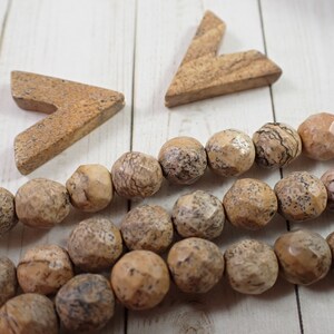 May include: A strand of brown and beige gemstone beads with a faceted finish. The beads are arranged in a loose pile on a white wooden surface. Two small, brown, triangular shapes are also on the surface.