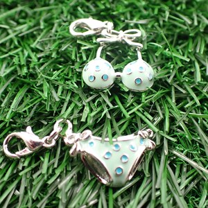 Könnte beinhalten: Miniatur-Bikini-Anhänger-Set aus silberfarbenem Metall mit hellblauem Emaille und türkisfarbenen Punkten. Das Set enthält ein Bikini-Oberteil und -Unterteil, jeweils mit einem Karabinerverschluss. Die Anhänger werden auf einem Bett aus grünem Kunstgras präsentiert.