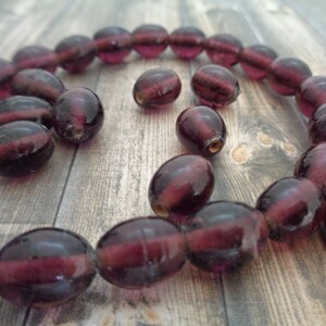 May include: A strand of deep purple glass beads, with a few extra beads scattered around. The beads are oval shaped and have a slightly iridescent finish.