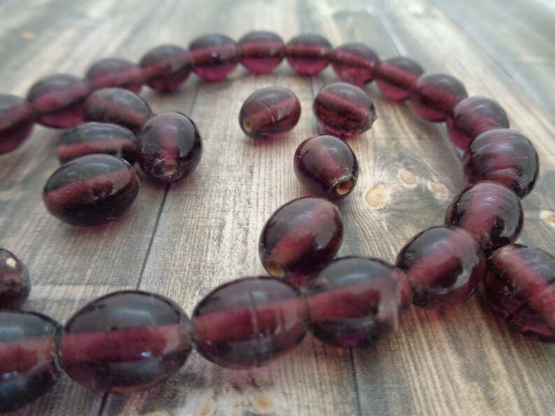 May include: A strand of deep purple glass beads, with a few extra beads scattered around. The beads are oval shaped and have a slightly iridescent finish.
