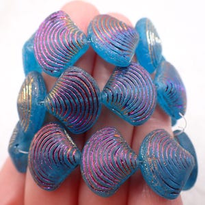 May include: A bracelet featuring multiple blue, shell-shaped beads with iridescent pink and gold accents. The beads are strung together, creating a unique and eye-catching accessory. The bracelet is held in a hand.