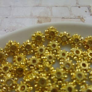 May include: A close-up of a pile of gold-colored metal beads. The beads are round with a raised, starburst design.