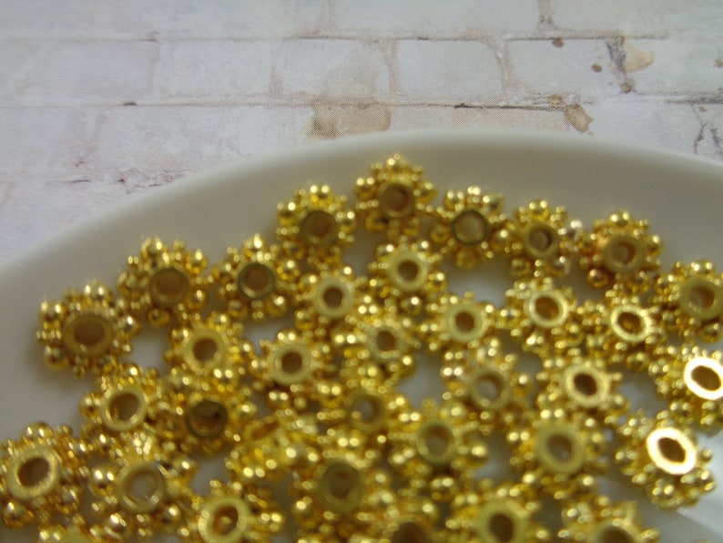 May include: A close-up of a pile of gold-colored metal beads. The beads are round with a raised, starburst design.
