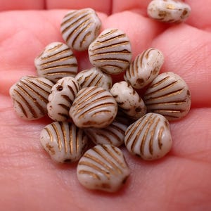 May include: A collection of off-white, heart-shaped beads with gold stripes. The beads are clustered together, showcasing their unique design and texture. These decorative beads are ideal for jewelry making and craft projects.