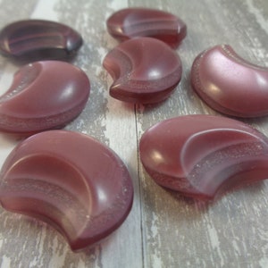 May include: Seven iridescent pink crescent moon shaped buttons with a textured surface.