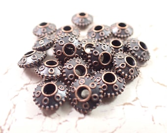 May include: A pile of 20 copper-toned metal beads with a decorative design. Each bead has a large hole in the center and a raised pattern of small circles around the edge.