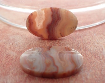 May include: Two oval-shaped agate cabochons with a banded pattern of white, brown, and orange. The cabochons are smooth and polished.