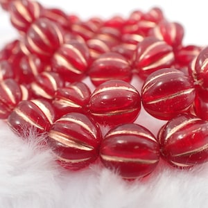 May include: Close-up of a strand of red glass beads with gold stripes. The beads are round and have a ribbed texture. The beads are resting on a white, fluffy surface.