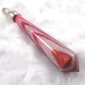May include: A pink and white crystal pendant with a silver-colored metal cap and a clear crystal loop. The pendant has a geometric shape with a pointed end and a marbled appearance.