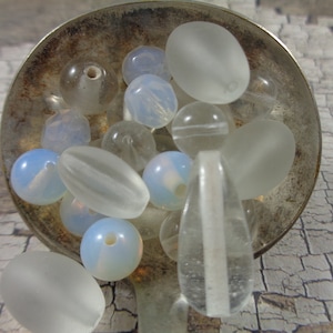 May include: A collection of white and blue glass beads in various shapes and sizes, including round, oval, and teardrop, displayed in a silver spoon.
