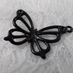 Op de afbeelding: Een zwarte metalen vlinderbedel met een uitgesneden ontwerp. De bedel heeft een lus aan de bovenkant om aan een ketting of armband te bevestigen.