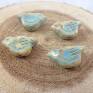 Op de afbeelding: Vier keramische vogel kralen in tinten van blauw en beige. De kralen zijn gevormd als vogels met vleugels en een kleine snavel. Ze zijn een geweldige aanvulling op elk juweelmaakproject.