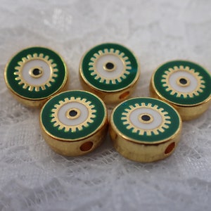 Op de afbeelding: Zes gouden kralen met een groen en wit emaille ontwerp. Elke kraal heeft een witte cirkel met een zwart oog in het midden, omringd door een gouden tandwielontwerp.