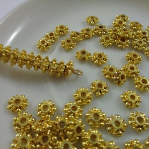 May include: A close-up of a pile of gold-colored metal beads with a flower-like design. The beads have a small hole in the center and are arranged in a random pattern.
