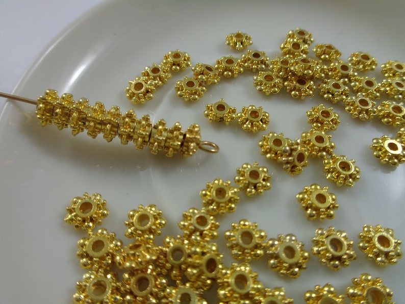 May include: A close-up of a pile of gold-colored metal beads with a flower-like design. The beads have a small hole in the center and are arranged in a random pattern.