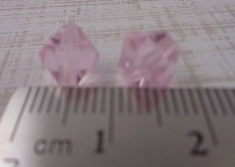 Puede incluir: Dos cuentas de cristal facetadas de color rosa, cada una mide aproximadamente 1 cm de di&aacute;metro, se muestran junto a una regla.