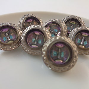 May include: Seven antique silver-coloured buttons are displayed. Each button features a detailed, textured border and an iridescent centre with purple, blue, and green hues. The buttons are arranged on a white surface.
