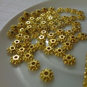 May include: A close-up of a pile of gold-colored metal beads with a starburst design. The beads are arranged on a white surface.