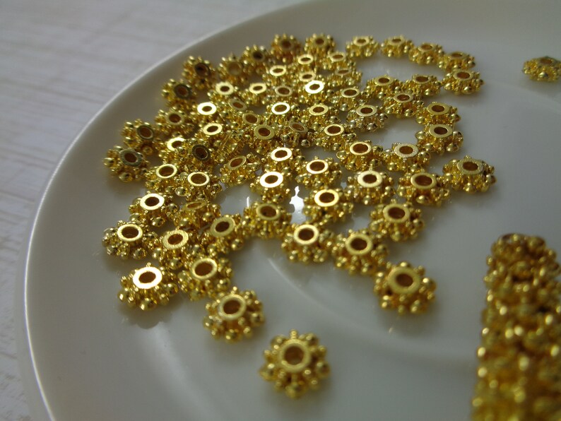 May include: A close-up of a pile of gold-colored metal beads with a starburst design. The beads are arranged on a white surface.