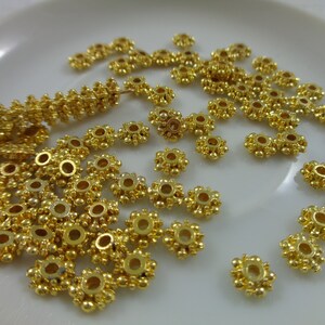 May include: A close-up of a pile of gold-colored metal beads. The beads are round with a small hole in the center and have a decorative design of small raised dots around the edge.