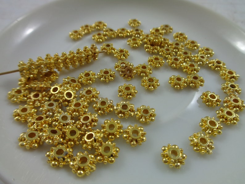 May include: A close-up of a pile of gold-colored metal beads. The beads are round with a small hole in the center and have a decorative design of small raised dots around the edge.