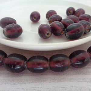 May include: A strand of deep purple glass beads, with a few loose beads on a white plate. The beads are oval shaped and have a translucent quality.