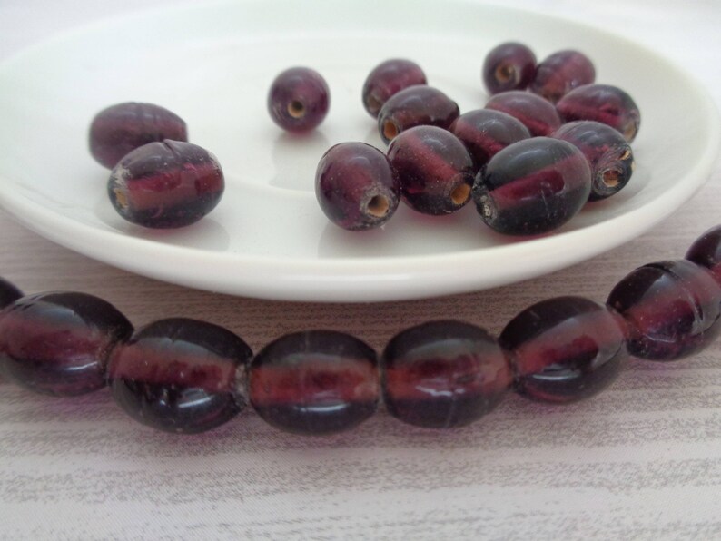 May include: A strand of deep purple glass beads, with a few loose beads on a white plate. The beads are oval shaped and have a translucent quality.