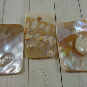 May include: Three rectangular pieces of iridescent mother-of-pearl shell with holes drilled in the top. The shell is a light beige colour with a pearlescent sheen.