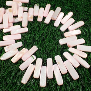 May include: A collection of pink, rectangular, shell beads with a pearly finish. The beads are arranged in a circle on a green, artificial grass surface.