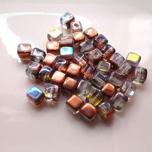 May include: A collection of iridescent cube-shaped beads in shades of copper, amber, and clear glass. The beads are scattered on a white surface, with some displaying a rainbow sheen. These beads are suitable for jewellery making and crafts.