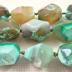 May include: Close-up of a strand of faceted agate beads in shades of green, brown, and yellow. The beads are strung on a green cord. The gemstones have a natural, organic appearance, with variations in colour and texture.