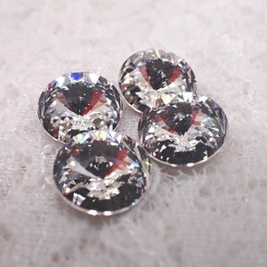 May include: Four clear, round rhinestones with multiple facets, reflecting light. The gems are arranged in a cluster, showcasing their brilliance. The background is a soft, white, textured fabric.
