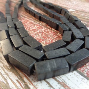 May include: A close-up of a black rectangular bead necklace. The beads are made of a smooth, polished material and have a slightly irregular shape. The necklace is laid out in a curved pattern on a wooden surface.