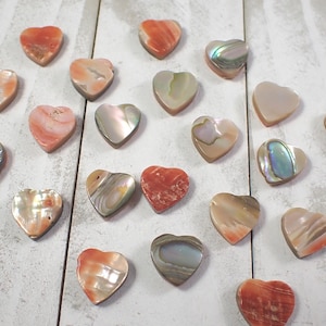 May include: A collection of heart-shaped stones in various colors and textures, including orange, pink, and iridescent mother-of-pearl. The stones are arranged on a white wooden surface.