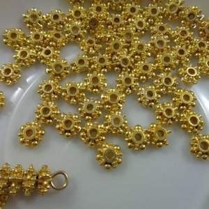 May include: A close-up of a pile of gold-colored metal beads with a flower-like design. The beads have a small hole in the center for stringing.