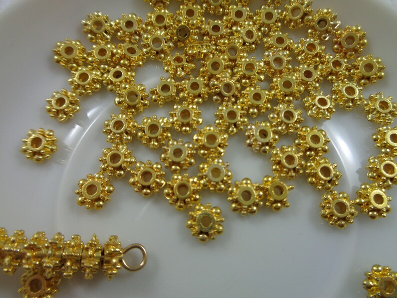 May include: A close-up of a pile of gold-colored metal beads with a flower-like design. The beads have a small hole in the center for stringing.