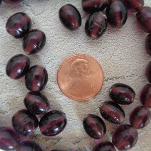 May include: A strand of dark purple glass beads, some of which are oval shaped and some are round. The beads are arranged in a circle around a penny.